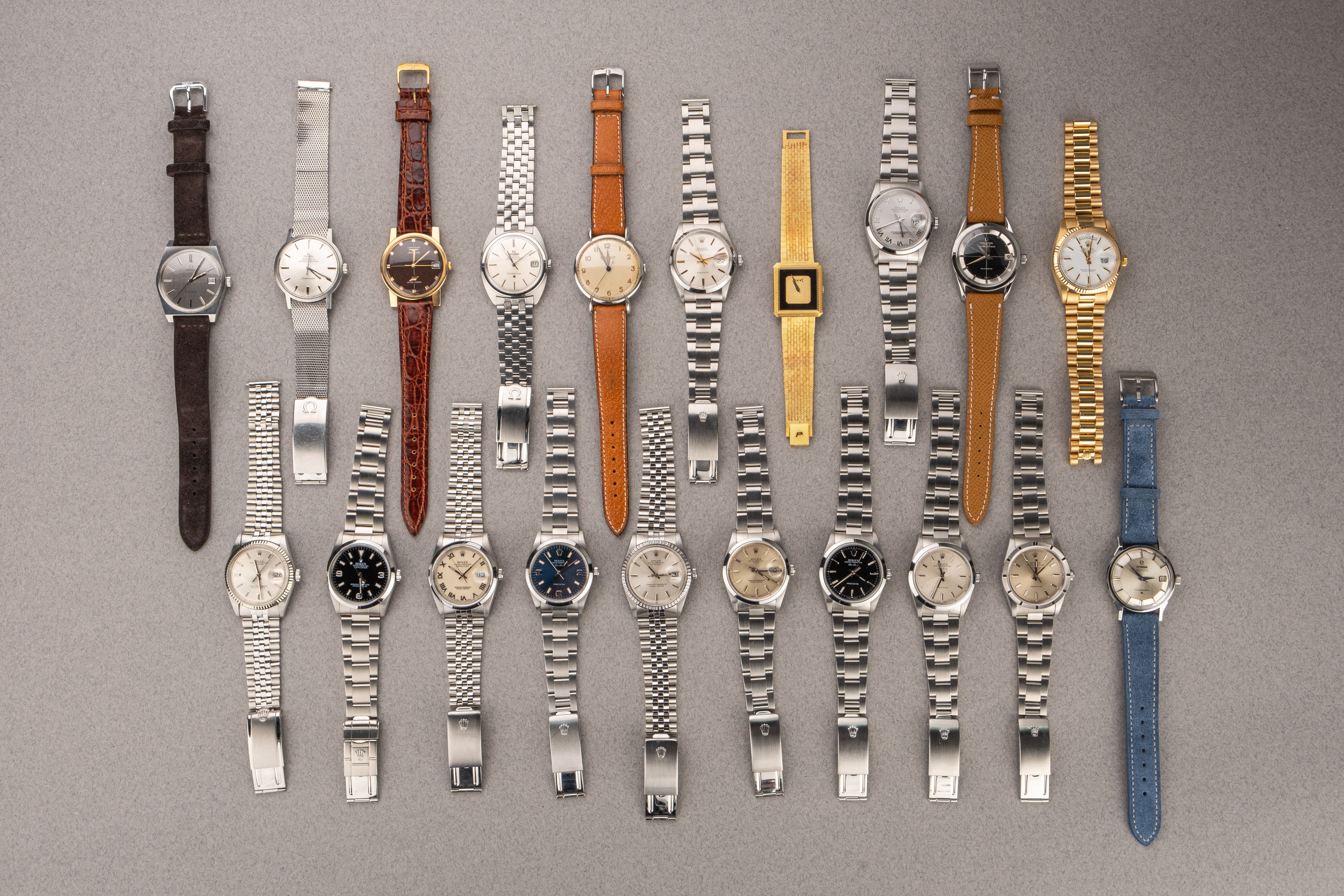 On a grey surface, many vintage watches by Rolex, Jaeger-LeCoultre, Omega, Longines, Piaget, Universal Genève and other Swiss brands for sale at Amplitude Vintage Watches in Paris, France.